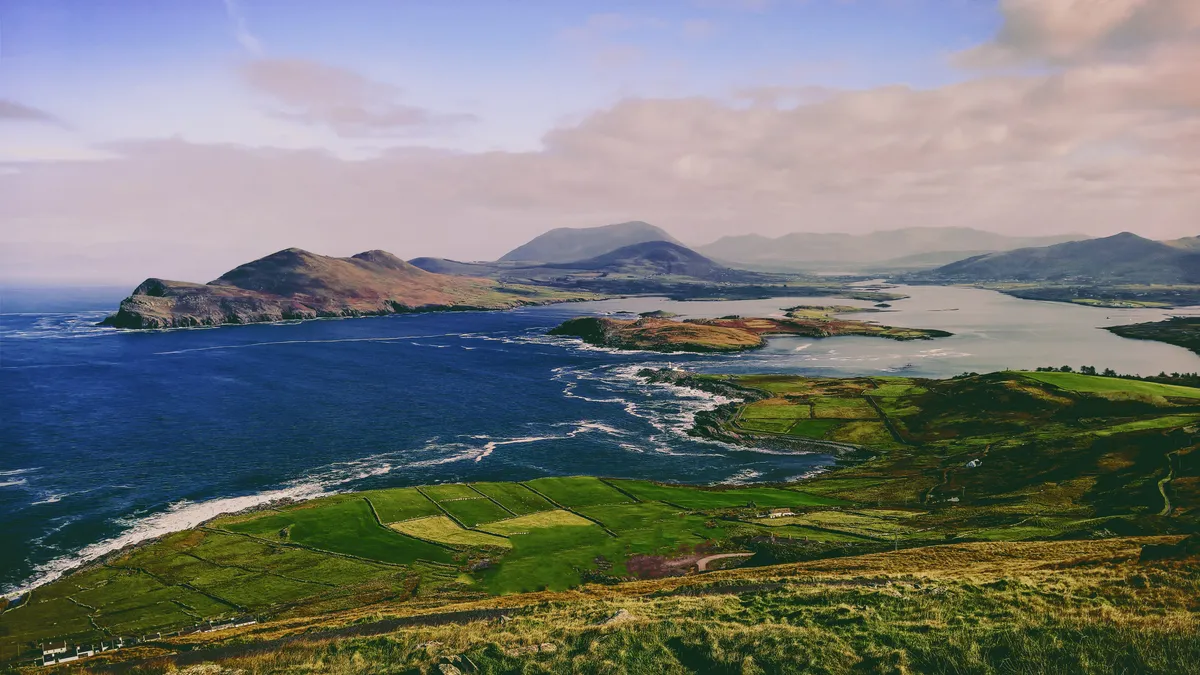 Aerial view of green coastal farmland on Valentia Island, County Kerry, Ireland, with the Atlantic beyond — infrastructure shaped by terrain.